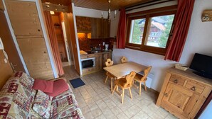 A cozy room with a wooden sofa, a dining table with chairs, a kitchen area with a stove, and a TV on a wooden cabinet. - 18m² Studio near slopes, balcony, parking (La Chapelle-d'Abondance)