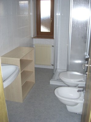 Baño con dos inodoros, un lavamanos, una estantería de madera, un radiador y una ventana.
