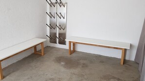 Zwei weiße Bänke mit Holzbeinen vor einer weißen Wand, im Hintergrund ein Metallgestell mit schwarzen Gegenständen.