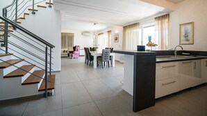 Una cucina moderna con isola centrale, zona pranzo con tavolo e sedie bianche, e una scala con gradini in legno e ringhiere in metallo.
