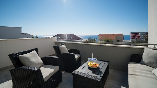 Un balcone con mobili in vimini, un tavolo con una ciotola di frutta e vista sul mare e sugli edifici.