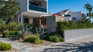 Una moderna casa a due piani con un balcone, immersa in un giardino curato con diverse piante e un vialetto lastricato.