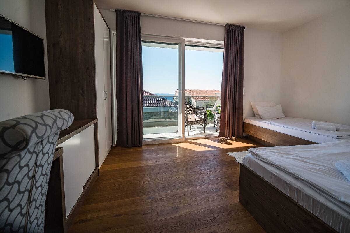 A hotel room with a balcony, wooden flooring, a bed, a sofa, and a TV.