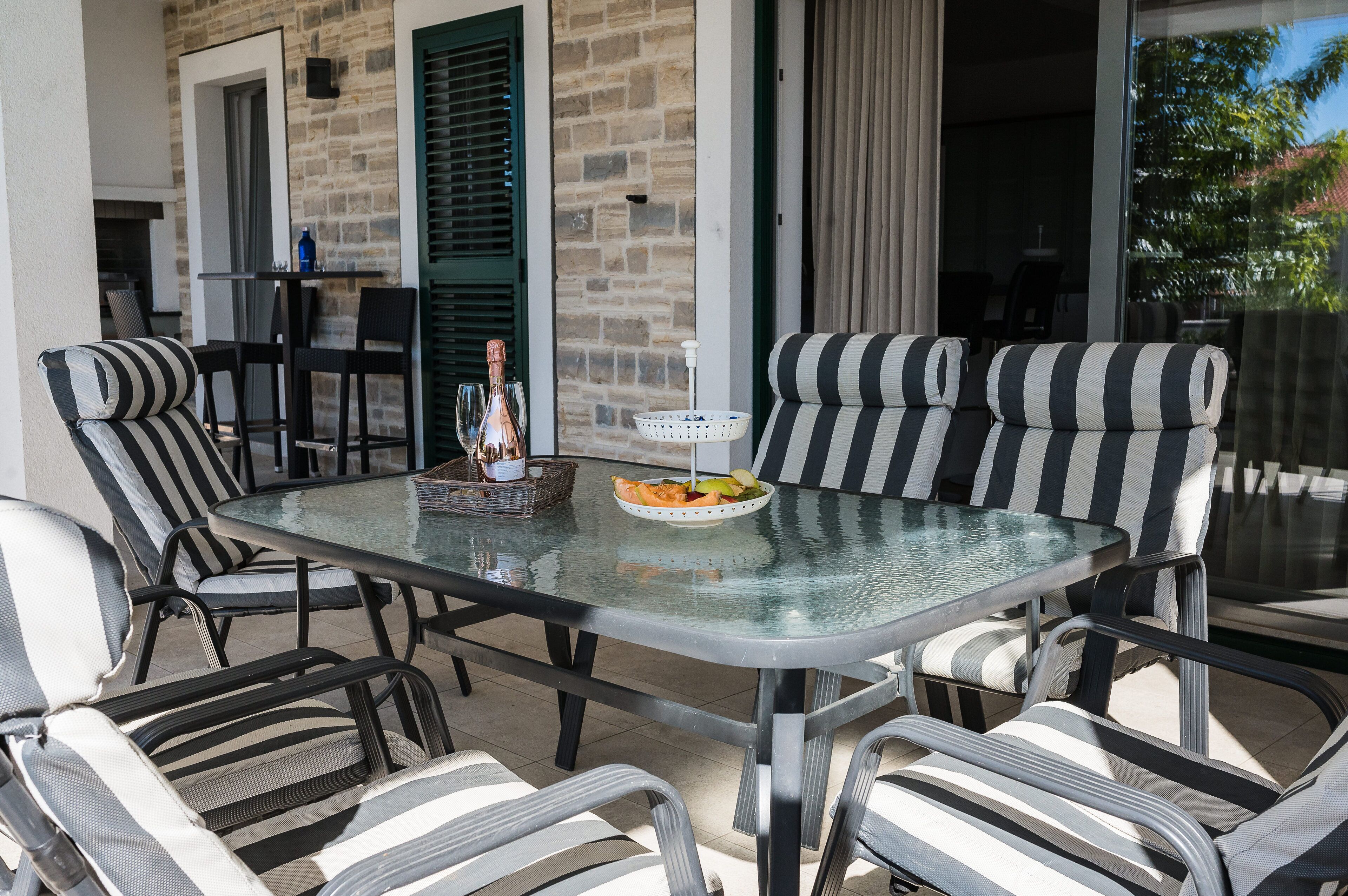 A glass-top table with striped chairs, a basket with fruits, and a bottle of wine.