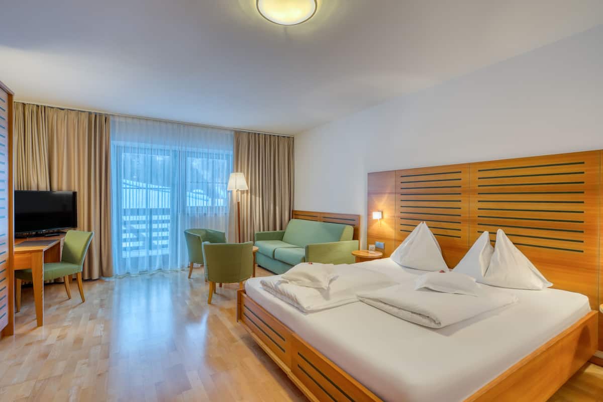 Habitación de hotel con cama de madera, un escritorio con televisor, dos sillas verdes, un sofá y un amplio ventanal con cortinas.