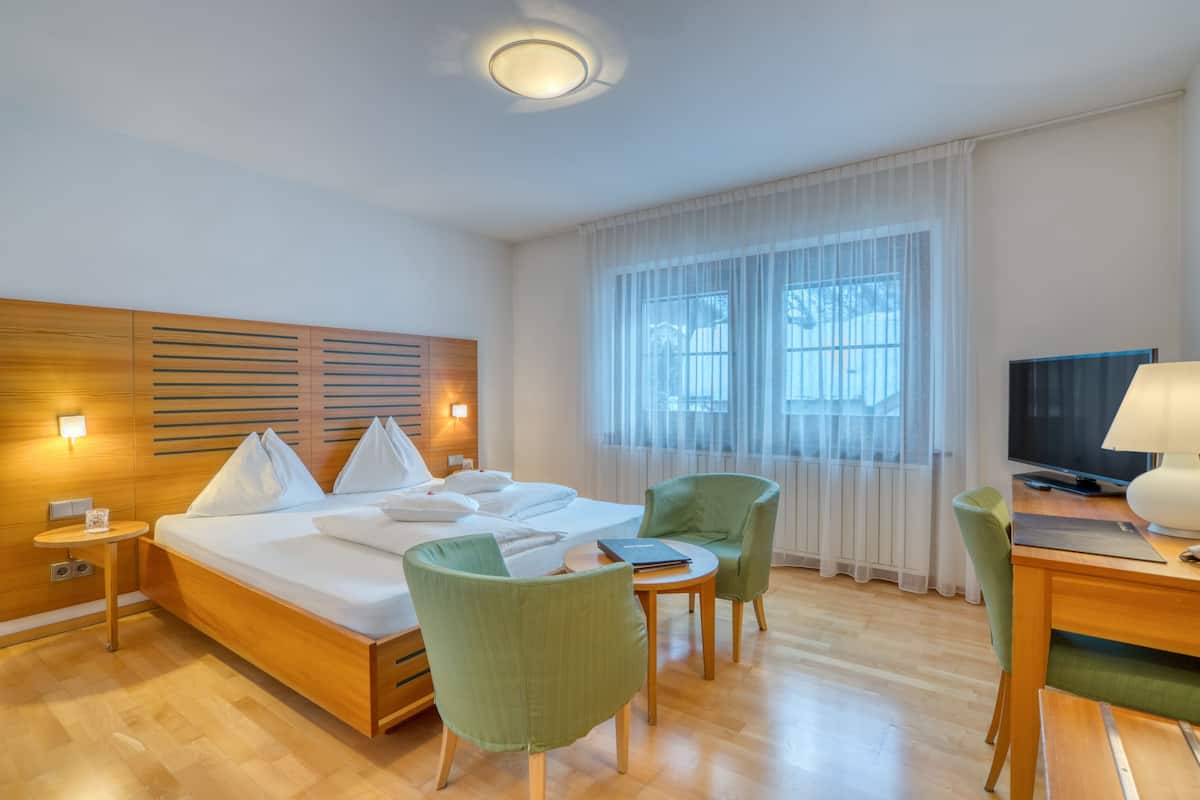 Habitación de hotel con una cama de madera, sillas verdes, un televisor y un escritorio con lámpara.
