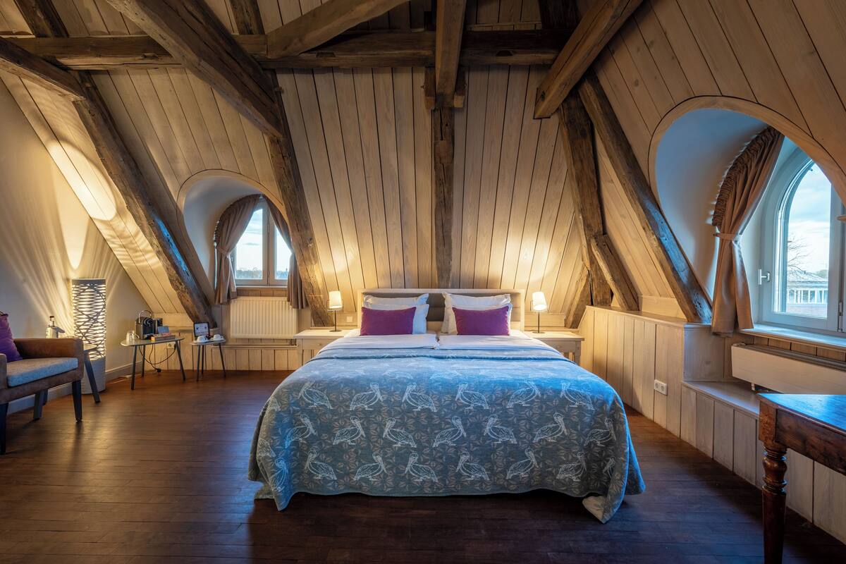 Une chambre avec un plafond en pente, des poutres en bois, un lit recouvert d’une couvre-lit à motifs, deux tables de chevet avec des lampes et une fenêtre avec des rideaux.