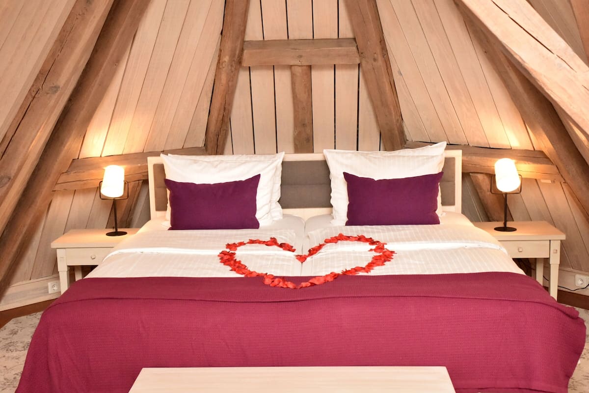 Une chambre mansardée chaleureuse avec un plafond en pente en bois, un lit avec une literie blanche et violette, et deux lampes de chevet.