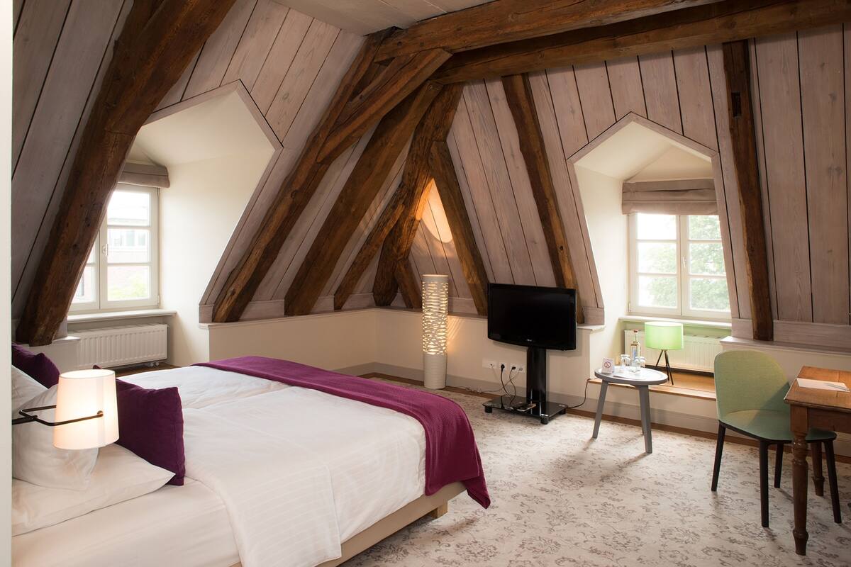 Une chambre avec un plafond en pente, un lit avec une literie blanche, une télévision, une lampe et une chaise.