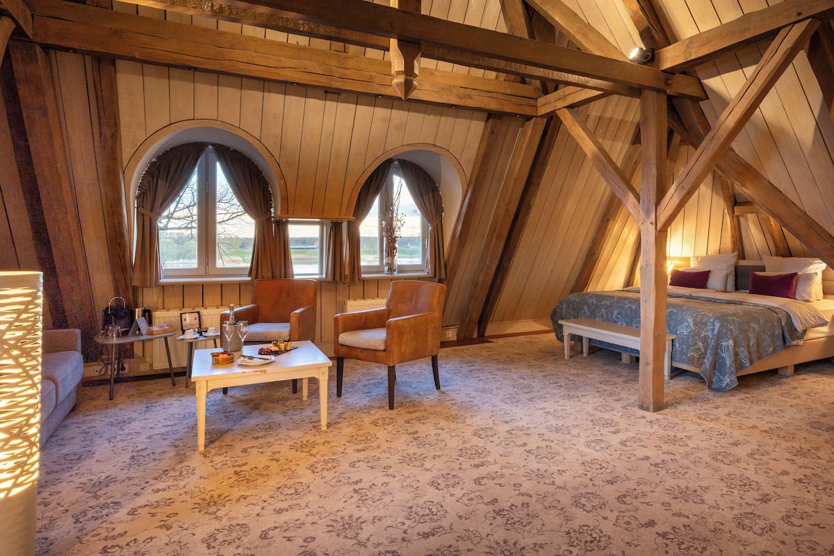 Une chambre confortable située dans les combles, avec un plafond en bois, deux fenêtres en arc de cercle, un lit recouvert d’une couverture bleue et une petite table avec une lampe.