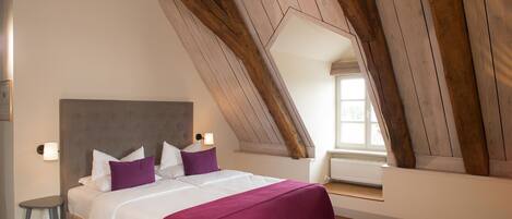 A bedroom with a sloped ceiling, a bed with white linens and purple pillows, a nightstand, and a window with natural light.