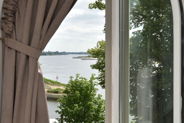 A view through a window with curtains, featuring a body of water, trees, and a distant shoreline.
