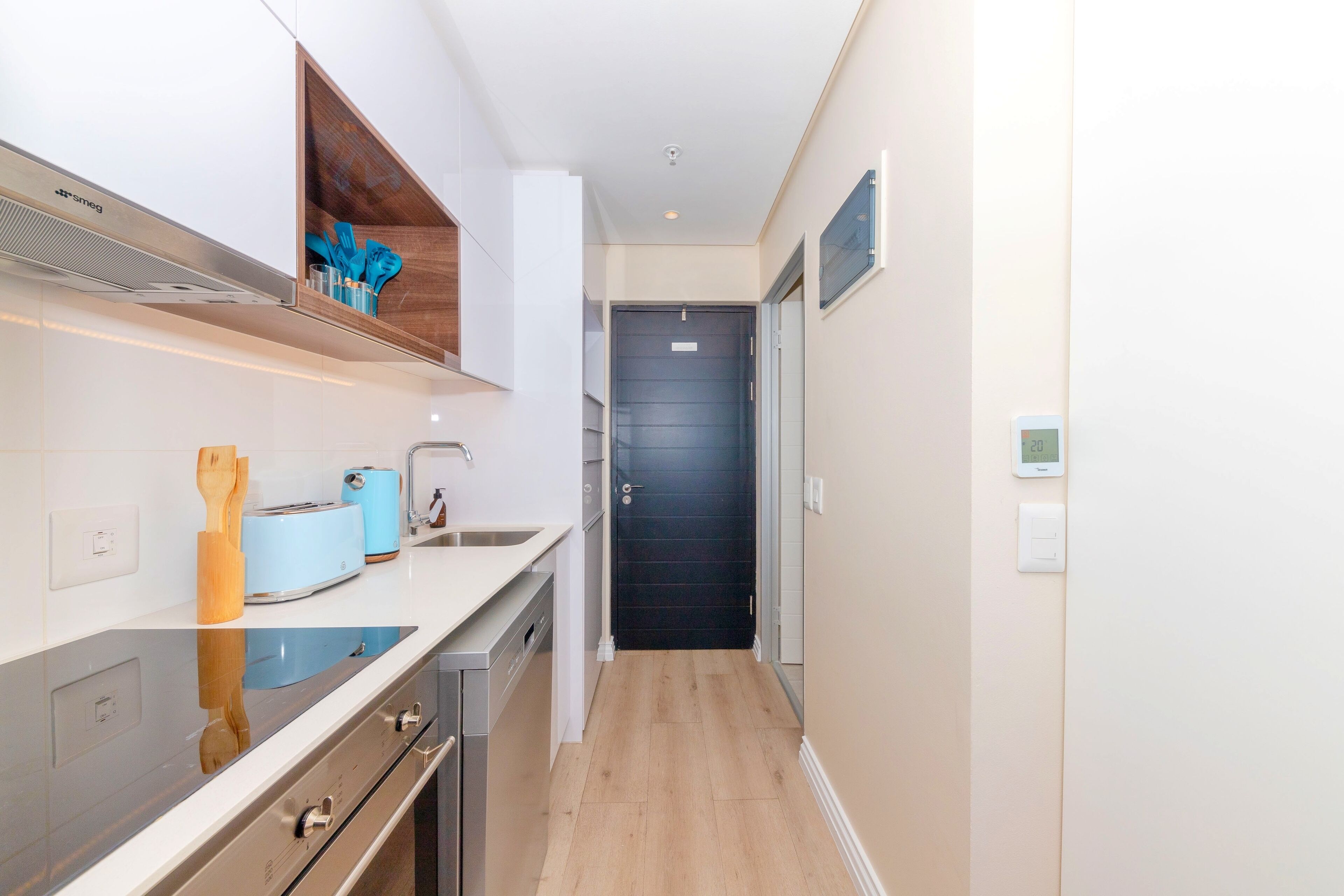 Ett smalt kök med modern design, utrustat med en inbyggd ugn, en diskho och en hylla i trä där köksredskap förvaras.
