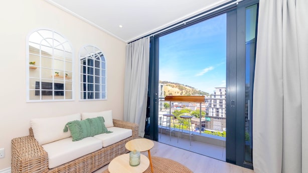 A modern living room with a wicker sofa, a small round table, and a large window offering a city view. - 16 on Bree 1218 (Cape Town)
