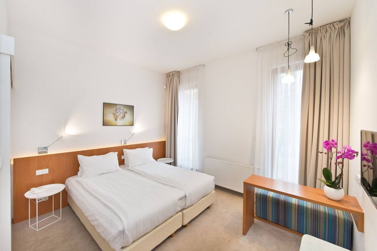 A modern hotel room with a large bed, bedside tables, a bench, and a potted plant.