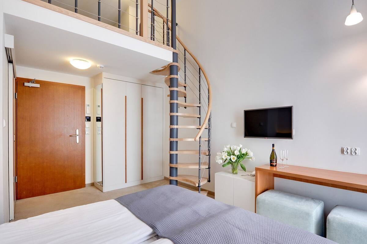 A modern hotel room with a bed, a desk with two chairs, a TV, a staircase, and a door.
