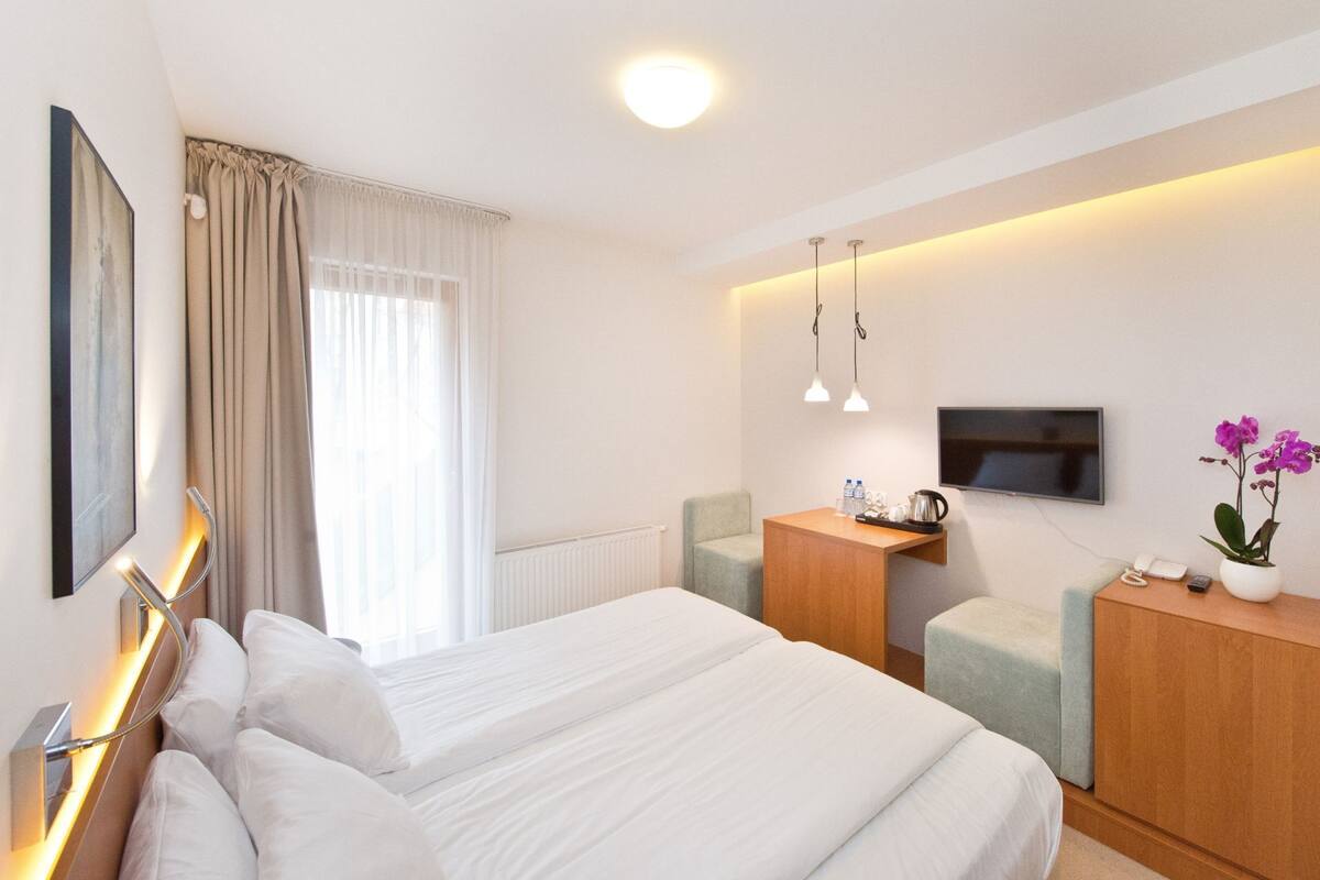 A modern hotel room with a large bed, a desk with a flat-screen TV, a chair, and a small table with a coffee maker.