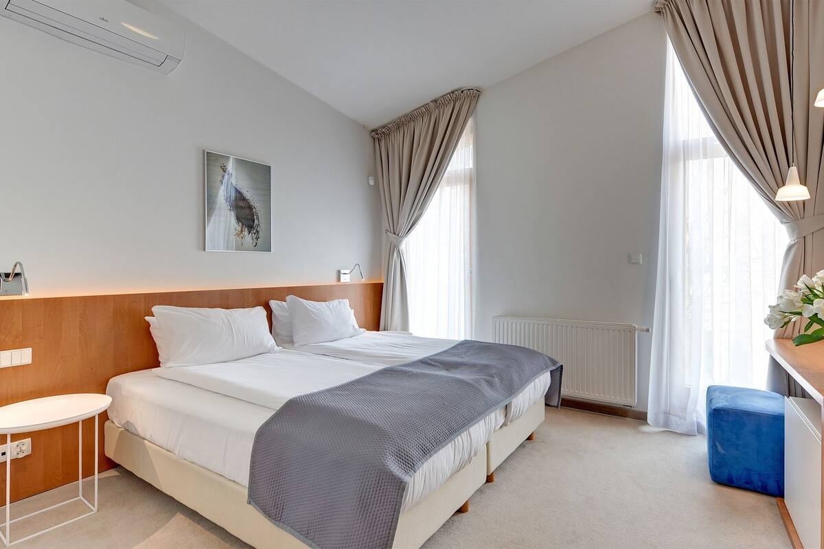 A hotel room with a large bed, a bedside table, a small stool, a desk with a chair, and a vase with flowers.