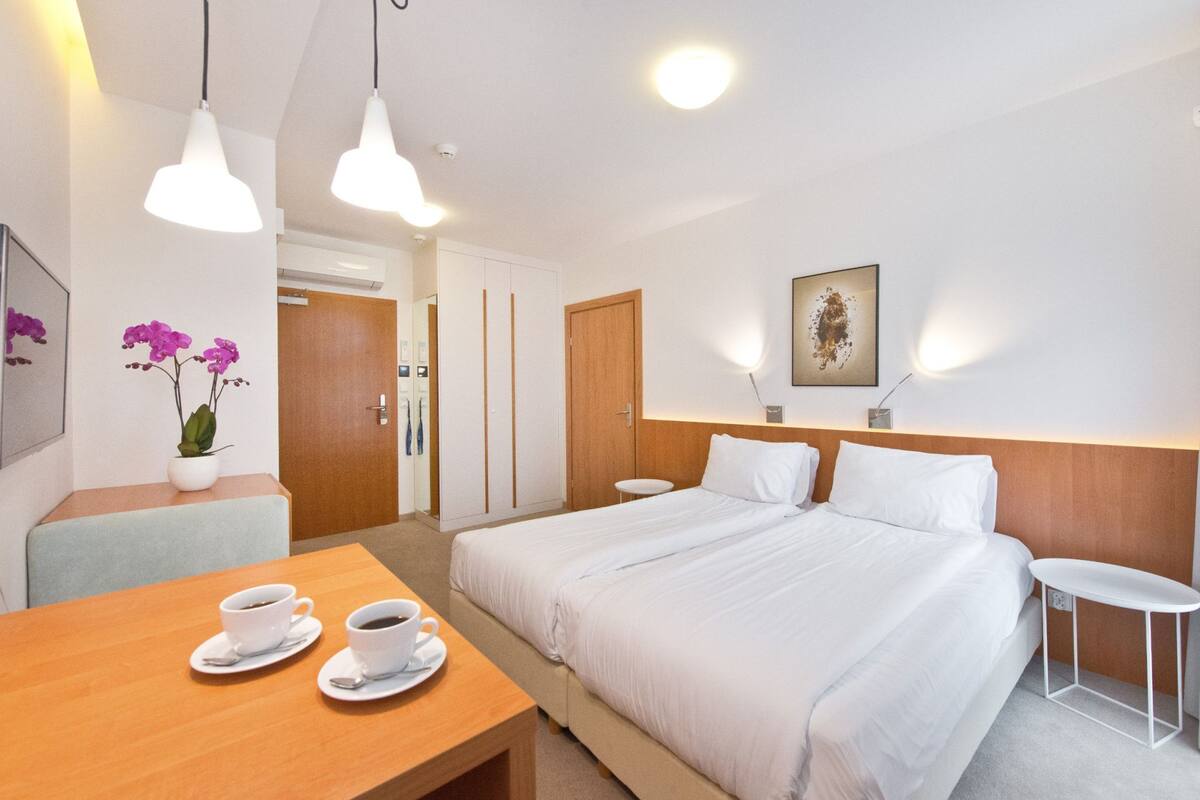 A hotel room with a bed, bedside tables, a desk with two cups of coffee, a potted orchid, and a painting on the wall.