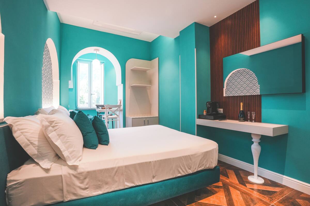 Une chambre moderne avec des murs de couleur turquoise, un lit avec une literie blanche et verte, un plancher en bois et une petite table sur laquelle se trouvent une bouteille et des verres.