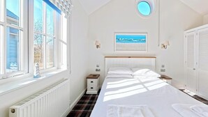 A bedroom with a bed, a nightstand, a wall-mounted lamp, a framed picture, and a window with blinds. - Stunning Beach House - Sleeping 4 in 2 Bedrooms - Pool and Beach Access Included. (Filey)