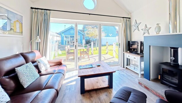 A living room with a sofa, coffee table, TV, fireplace, and a view of a blue house through large glass doors. - Stunning Beach House - Sleeping 4 in 2 Bedrooms - Pool and Beach Access Included. (Filey)