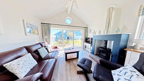 A living room with a brown leather sofa, a wooden coffee table, and a black armchair. There is a fireplace with a black stove, a television, and a dining area visible through large glass doors. - Stunning Beach House - Sleeping 4 in 2 Bedrooms - Pool and Beach Access Included. (Filey)