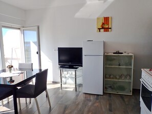 Ett kompakt kök med kylskåp, ugn och ett glasskåp med porslin. Där finns ett matbord med stolar och en tv på ett tv-bord.