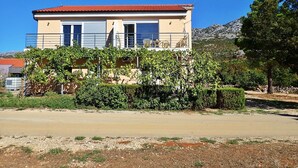 A two-story building with a red-tiled roof, a balcony with outdoor furniture, and a mountainous backdrop.