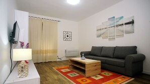 Un salon avec un canapé gris, une table basse en bois, une table d’appoint blanche avec une lampe, un tapis rouge et orange, et un téléviseur fixé au mur.