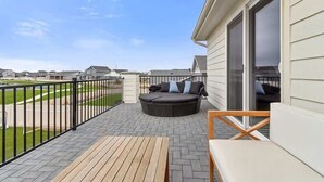 Une terrasse couverte avec une table en bois, un canapé d’extérieur noir avec des coussins et une vue sur un champ verdoyant.