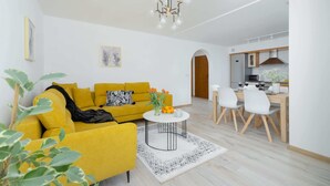 A modern living room with a yellow sofa, a round coffee table, and a dining area with white chairs. - Stylish Apartment with Balcony | Close to Mountain Trails (Zakopane)