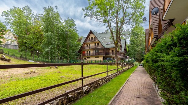 A traditional chalet-style building with a sloped roof, surrounded by lush greenery and a paved pathway. - Stylish Apartment with Balcony | Close to Mountain Trails (Zakopane)