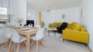 A modern living room with a yellow sofa, white chairs, a wooden dining table, and a chandelier. - Stylish Apartment with Balcony | Close to Mountain Trails (Zakopane)
