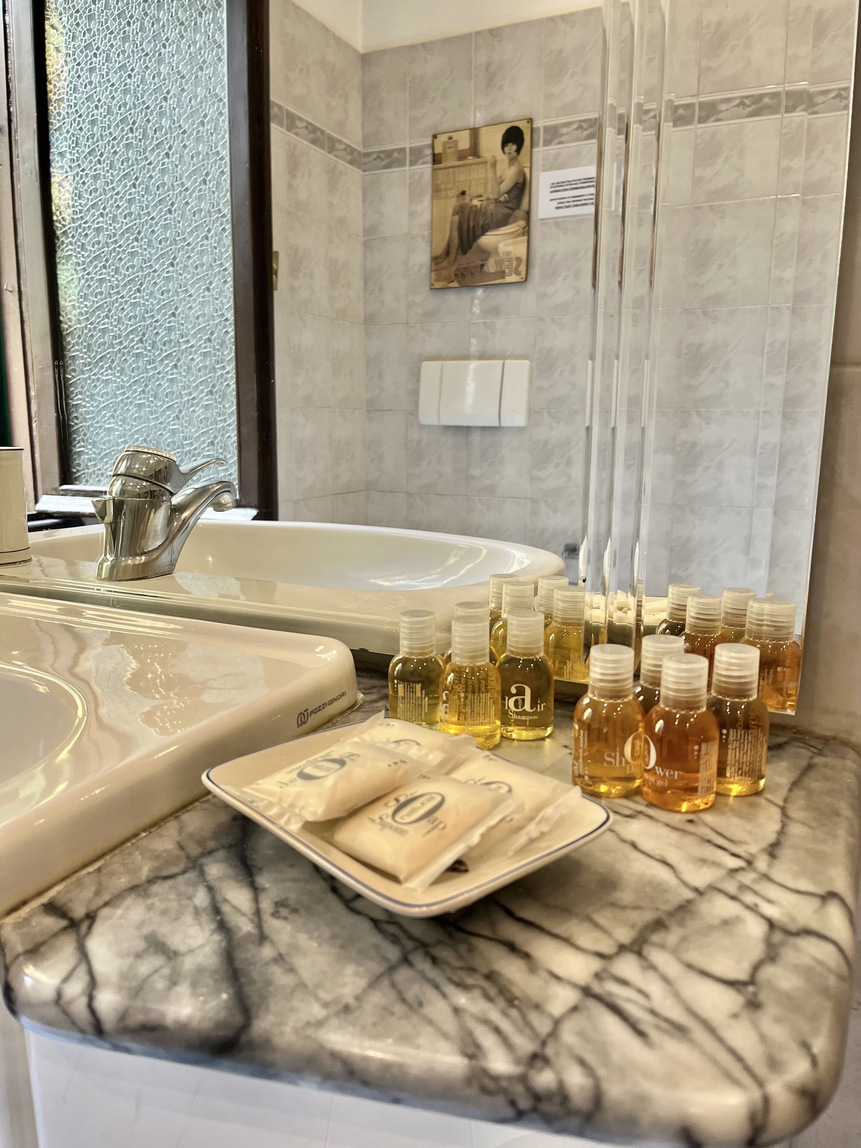 A bathroom with a marble countertop, a sink, and a shower area. There are several bottles of shampoo and conditioner on the counter.