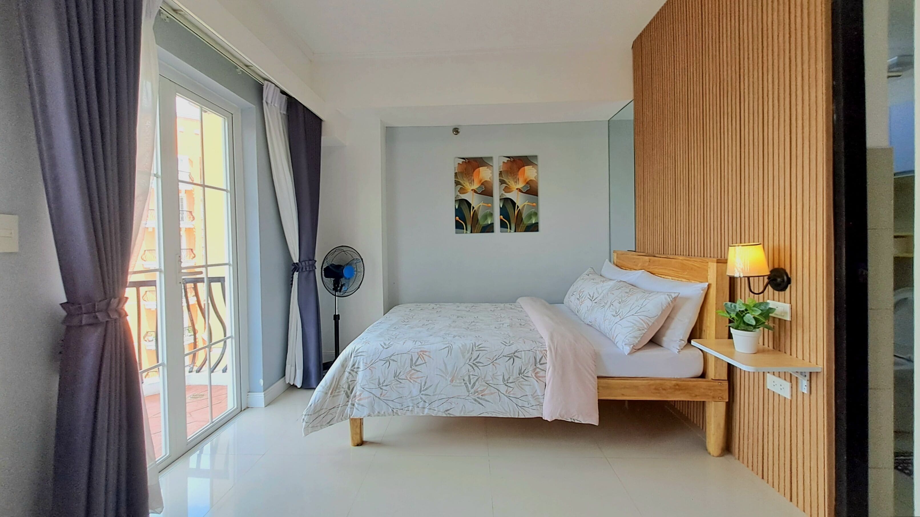 Ein Schlafzimmer mit Bett, Standventilator, Wandlampe und einem Fenster mit Vorhängen.