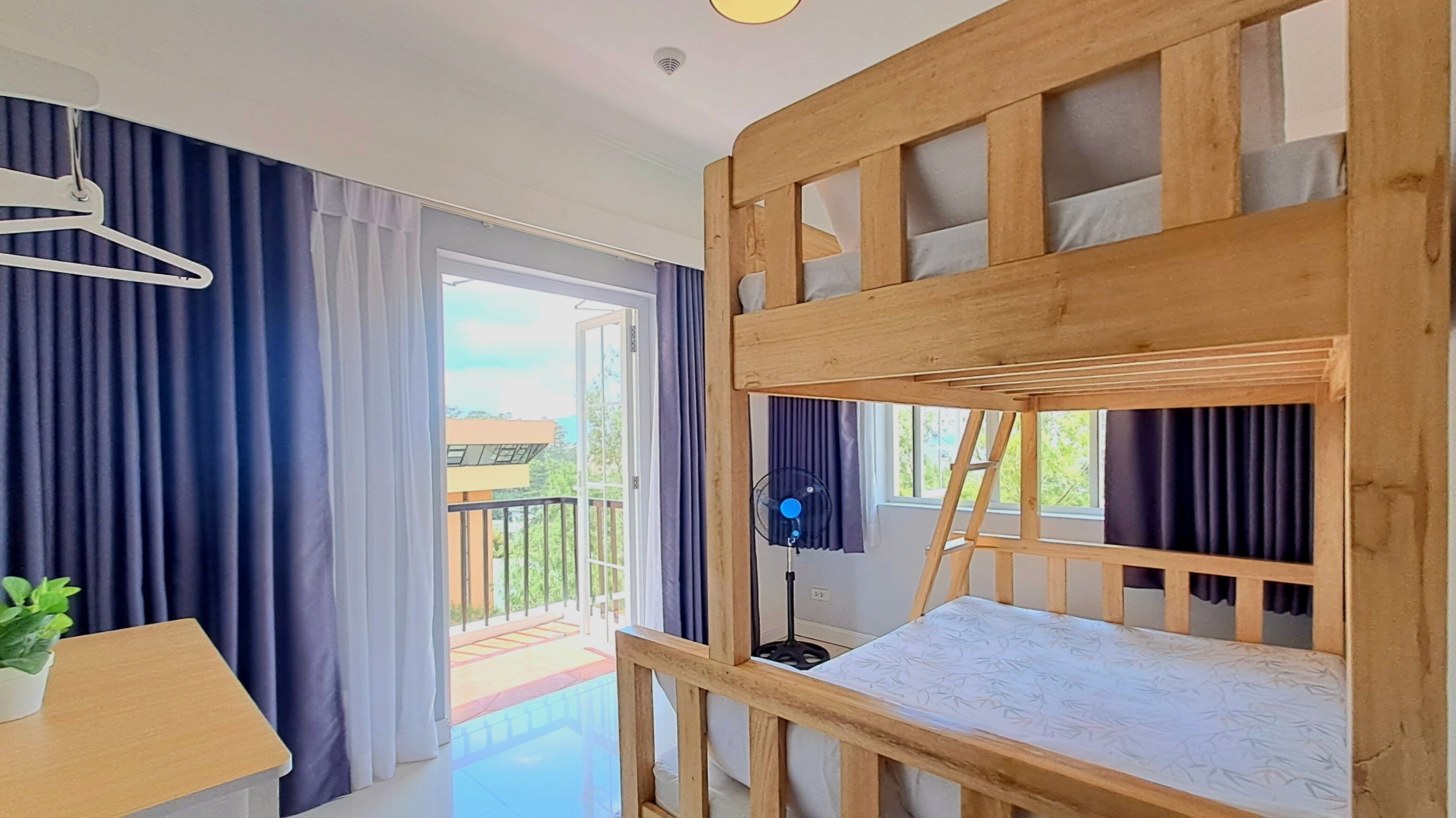 A room with a bunk bed, a desk with a plant, a fan, and a view of the outdoors.
