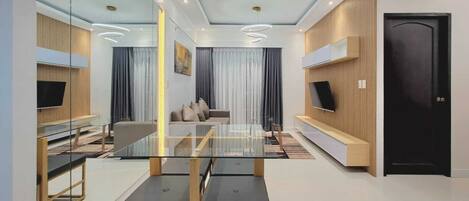 A modern living room with a glass dining table, sofas, and a TV mounted on the wall.