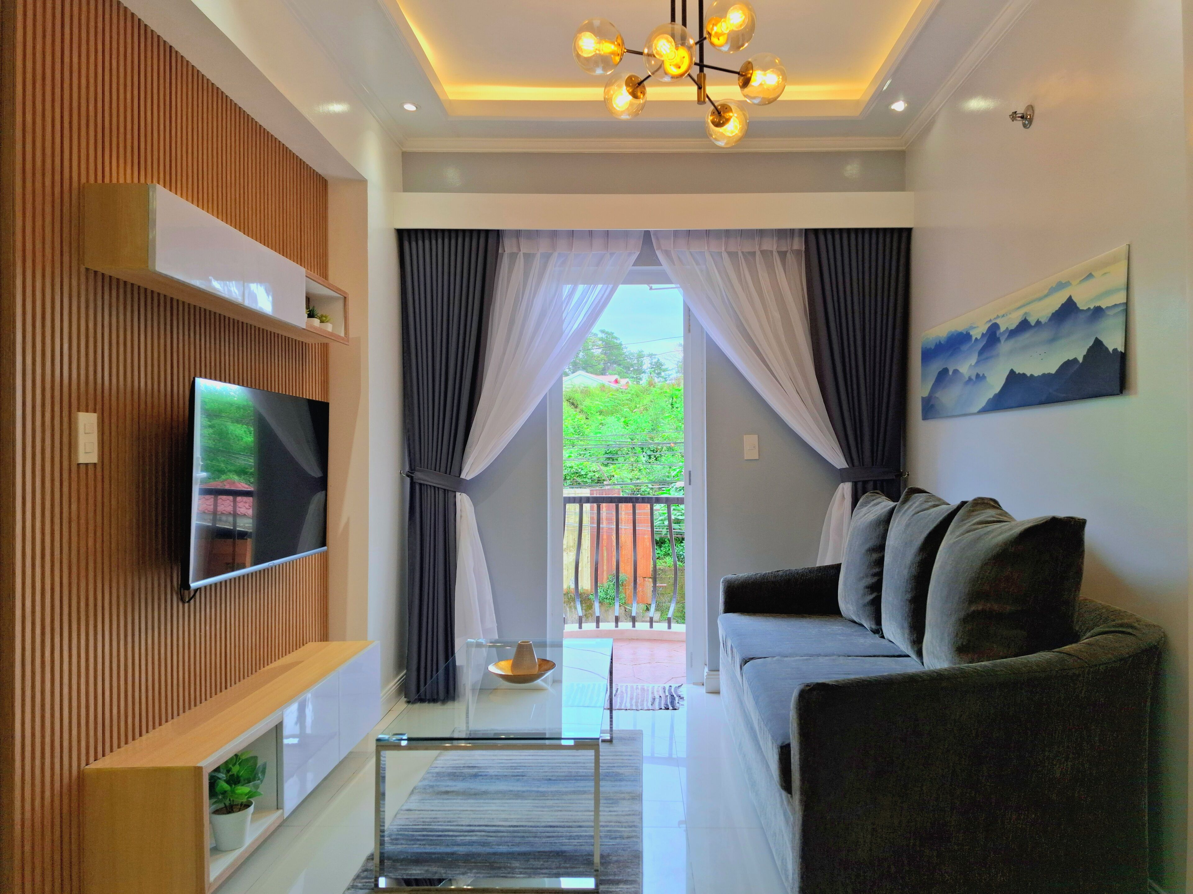 A modern living room with a flat-screen TV mounted on a wooden panel wall, a gray sofa, a glass coffee table, and a view of a balcony with greenery.