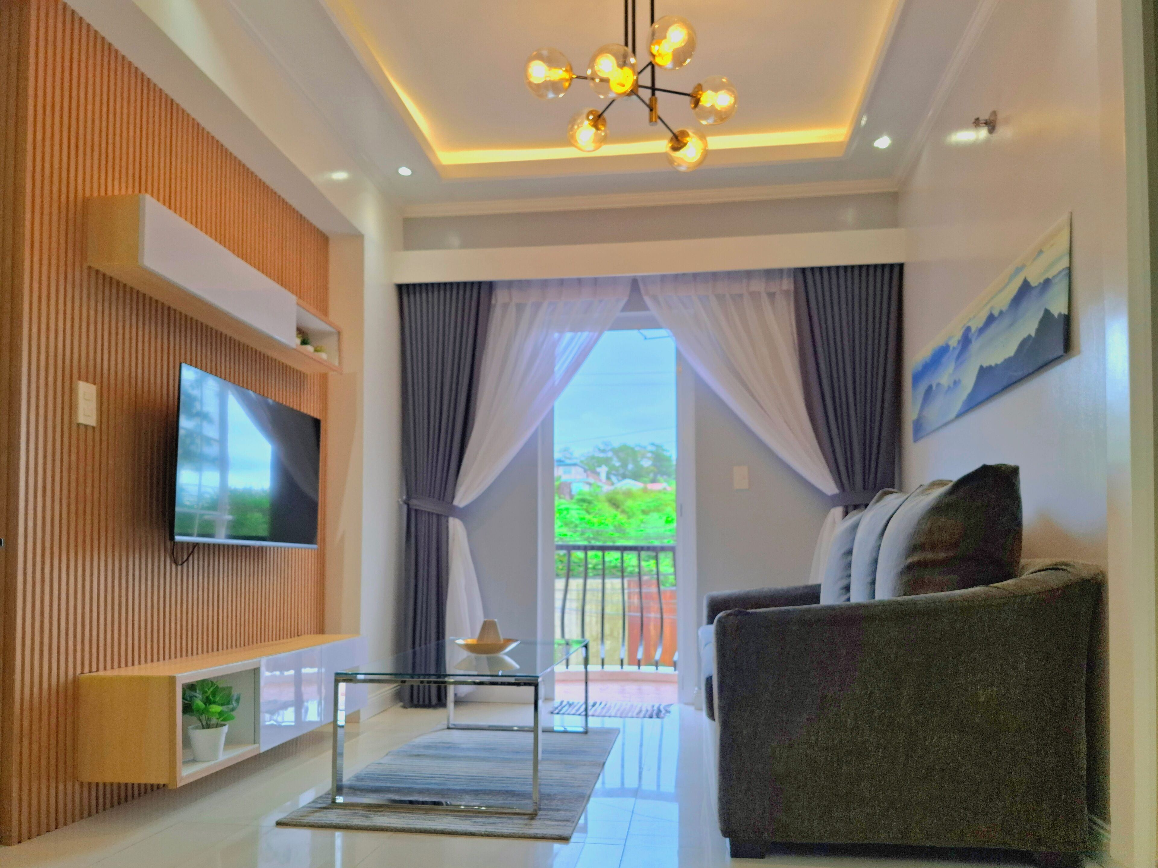 A modern living room with a flat-screen TV mounted on a wooden panel wall, a glass coffee table, a sofa, and a large window with curtains.