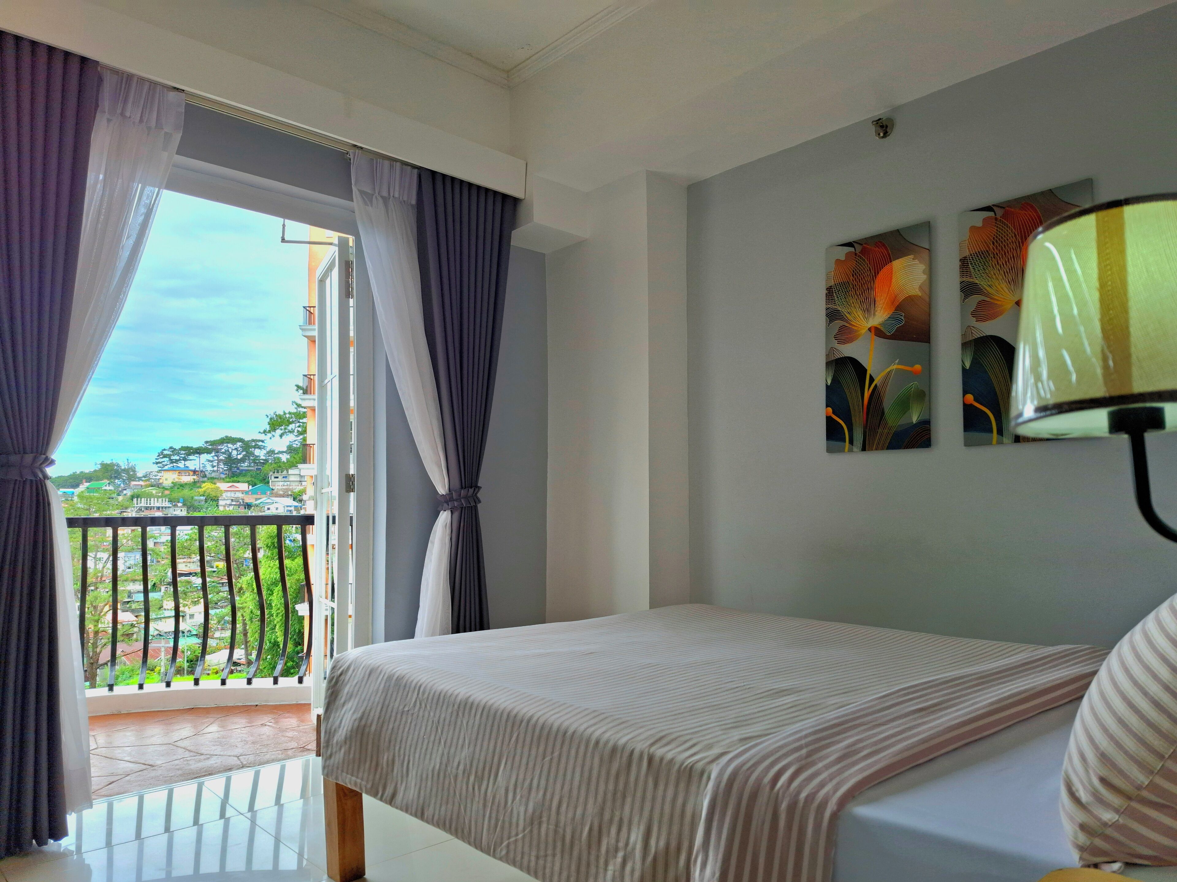 Habitación de hotel con cama, balcón con cortinas y un cuadro en la pared.