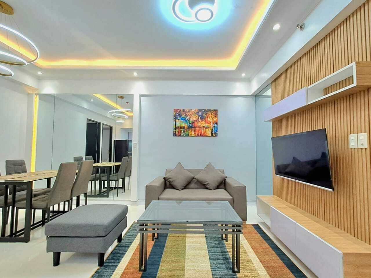 A modern living room with a sofa, coffee table, and TV. There is a dining area with chairs and a table in the background.
