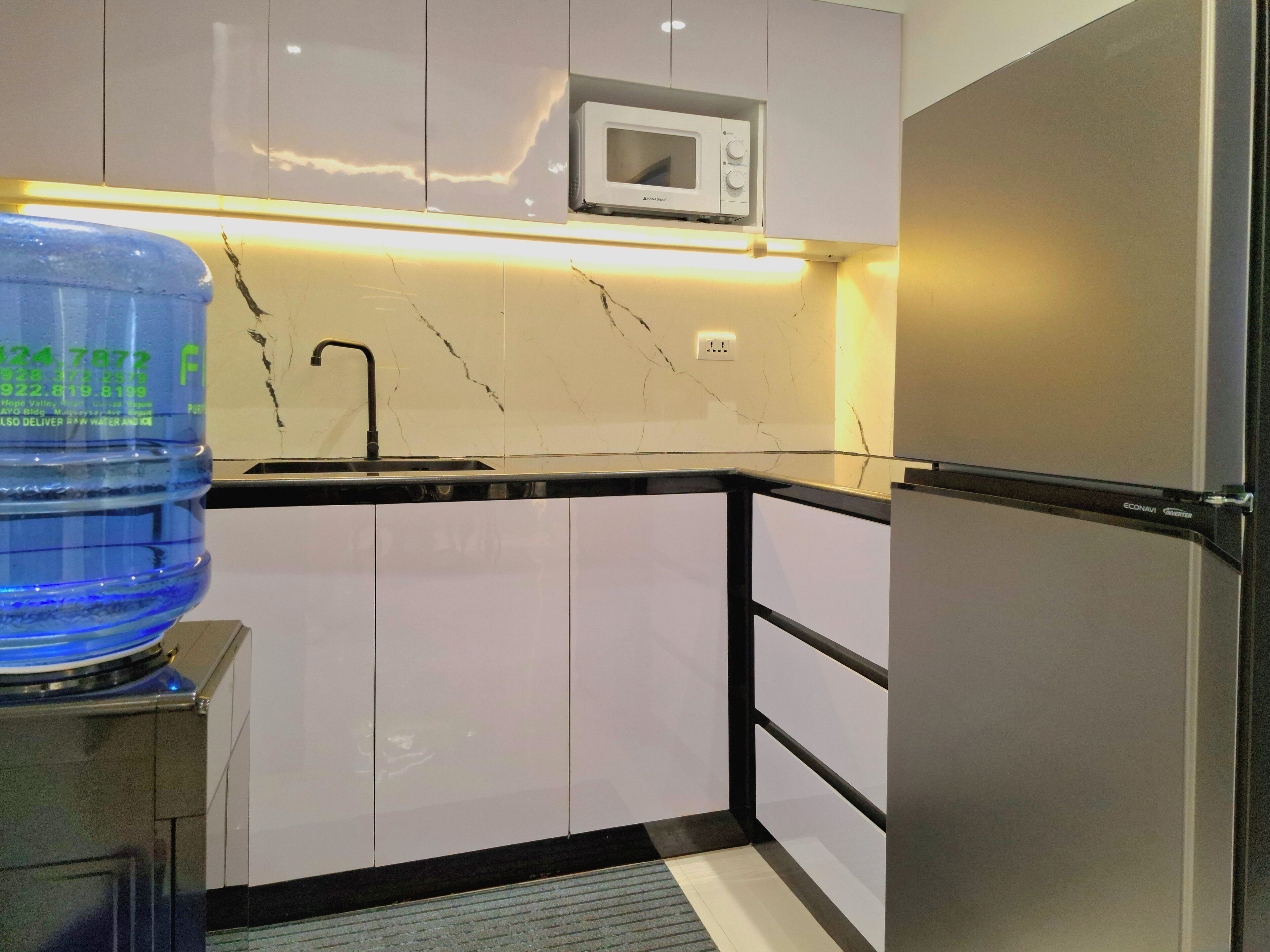 A compact kitchen with white cabinets, a black countertop, a built-in microwave, a water dispenser, and a refrigerator.