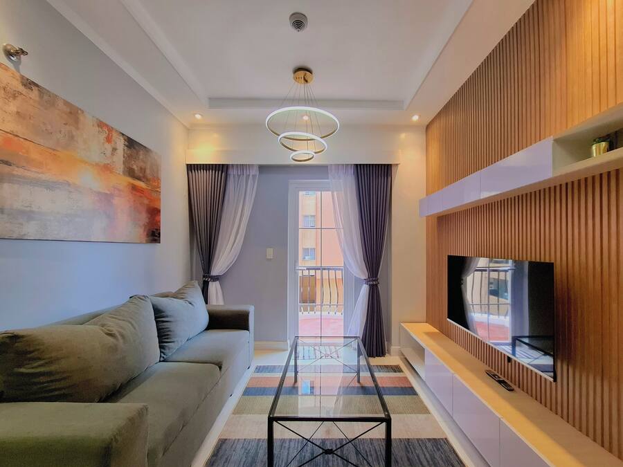 Een moderne woonkamer met een bank, een glazen salontafel, een tv die aan een houten paneel bevestigd is, en een groot abstract schilderij aan de muur.