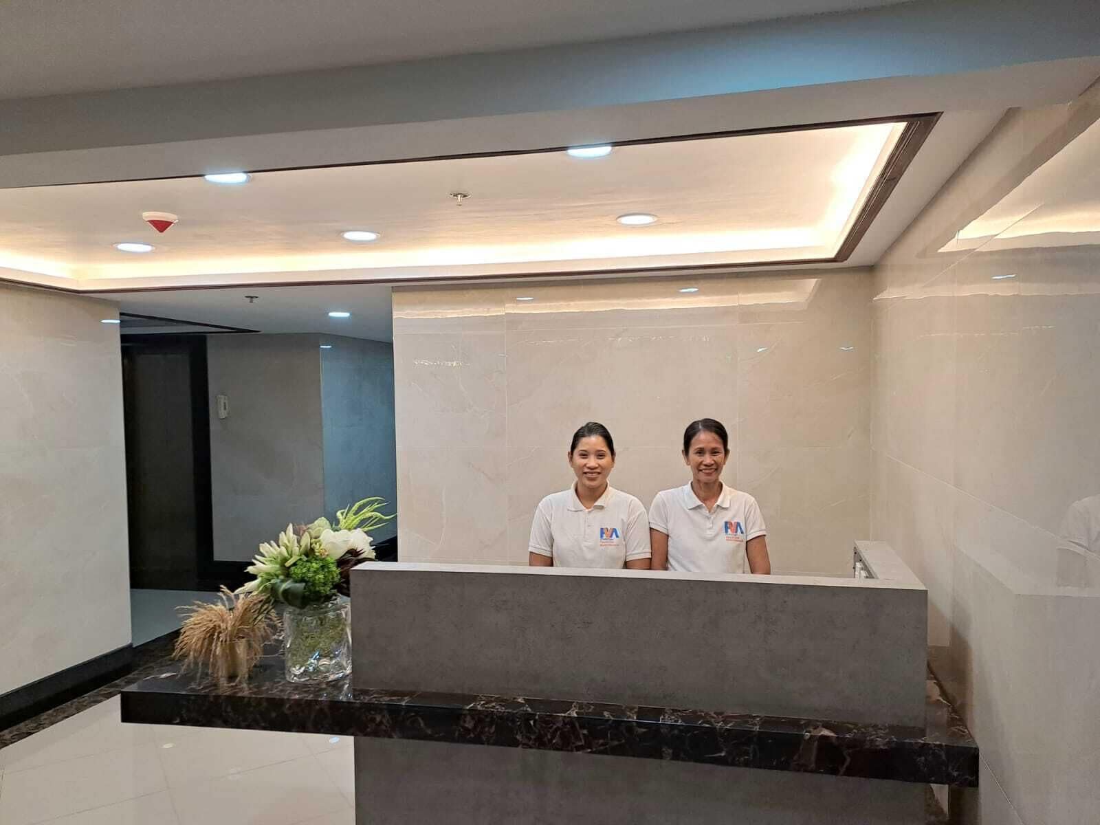 A reception area with a marble counter, two staff members in white uniforms, and a modern ceiling design.