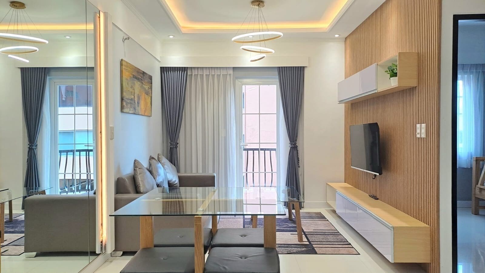 A modern living room with a glass dining table, a sofa, a TV mounted on a wooden panel, and a balcony with curtains.