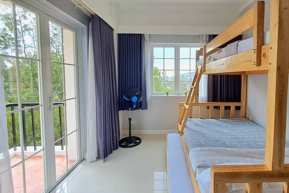 Een kamer met een stapelbed, grote ramen, een ventilator en gordijnen.