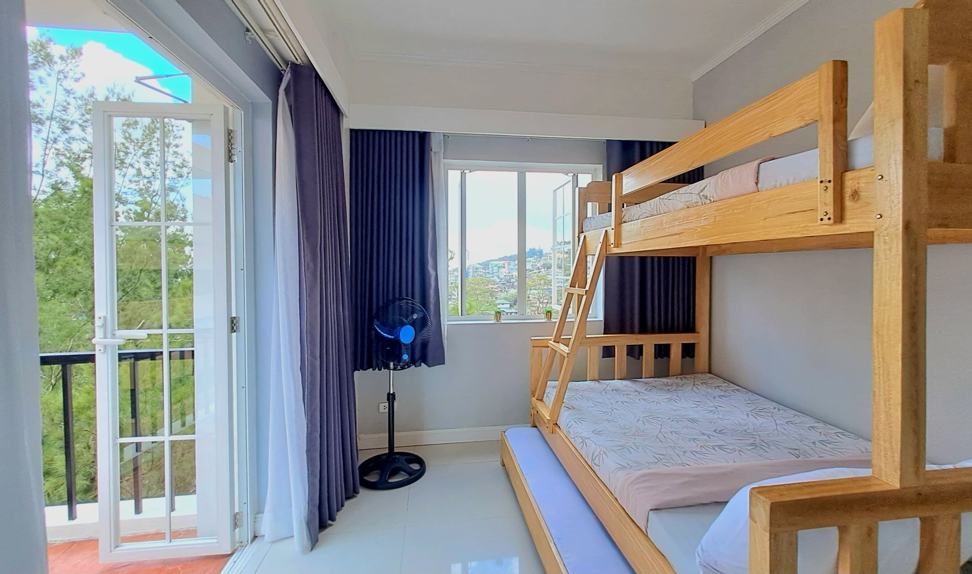 Ein Zimmer mit Etagenbett, großen Fenstern, einem Ventilator und Blick ins Freie.