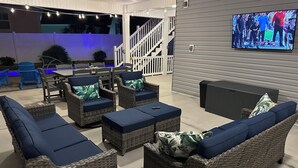 Een terras buiten met wicker meubels en een televisiescherm waarop een sportevenement wordt getoond.