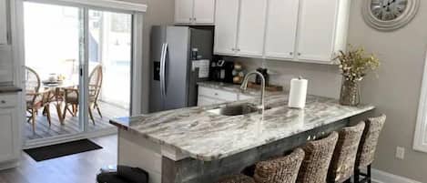 Una cocina con armarios blancos, una isla de mármol, taburetes de mimbre y un reloj en la pared.